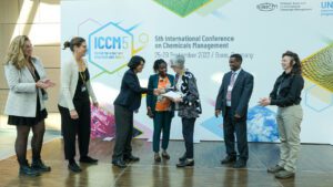 A group of seven smiling individuals stands in front of a banner for the ICCM5 conference as they deliver a signed petition to the Conference President who is standing near the center of the group. She is shaking hands with the individual who handed the petition to her.