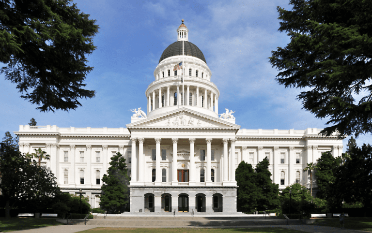 Image of the California Capitol during the day in front of a blue sky and framed by trees.